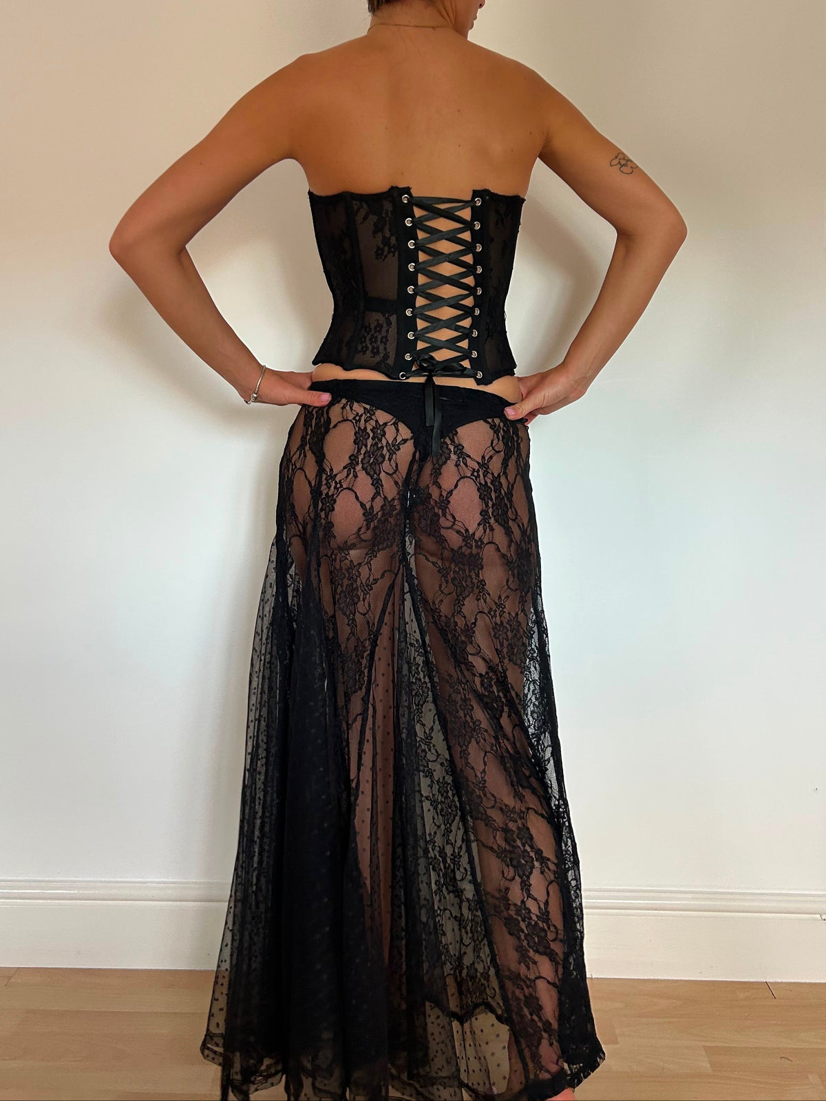 A woman stands with her back to the camera wearing a strapless black lace corset-style dress with a sheer tulle skirt, photographed indoors against a plain light wall.